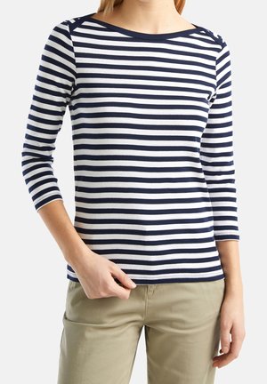Femme portant un haut à manches longues à rayures horizontales bleu marine et blanche et un pantalon beige, tenant l'ourlet du haut d'une main.