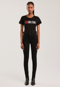 Tânără cu păr împletit, purtând un tricou negru "BIG STAR", pantaloni negri cu talie înaltă și cizme negre până la gleznă, stând în fața unui fundal simplu.