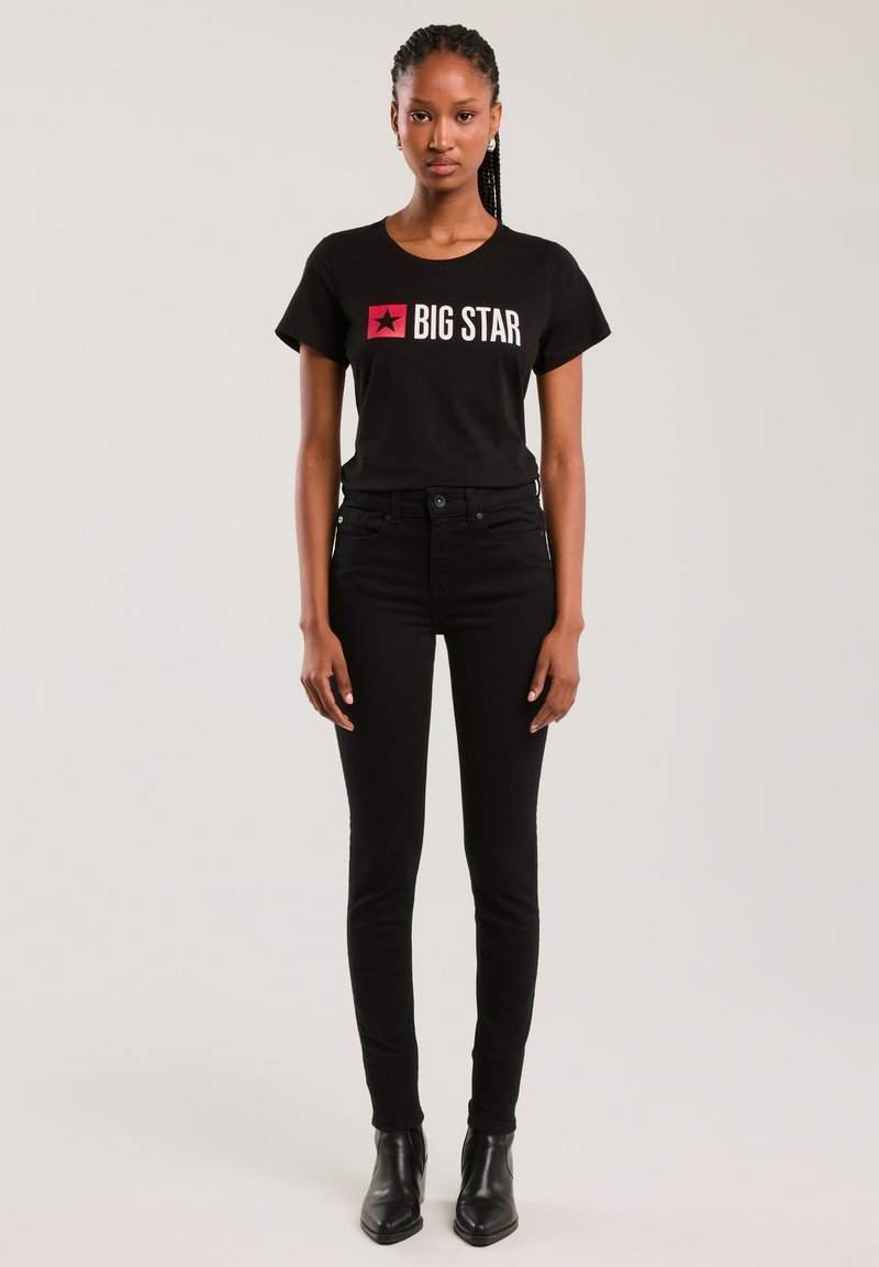 Tânără cu păr împletit, purtând un tricou negru "BIG STAR", pantaloni negri cu talie înaltă și cizme negre până la gleznă, stând în fața unui fundal simplu.