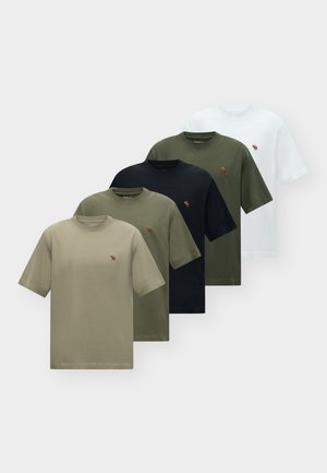 Jeune homme aux cheveux bouclés portant un t-shirt blanc, un short beige, une veste beige nouée autour de la taille, des chaussettes blanches et des baskets blanches, debout sur un fond blanc.