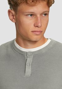 Graues, gestricktes Henley-Shirt mit strukturiertem Gewebe, das einen Rundhalsausschnitt und eine Knopfleiste mit zwei Knöpfen aufweist, getragen über einem weißen Hemd.