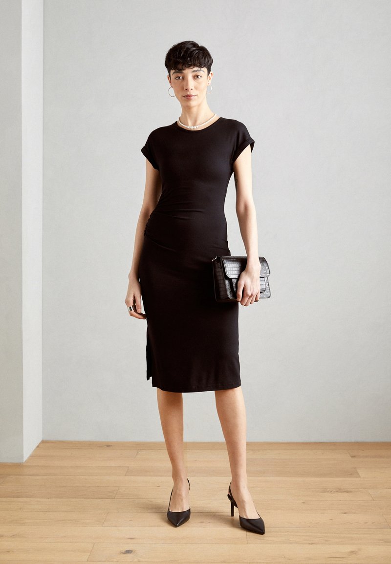 Czarna dopasowana sukienka z krótkimi, podwiniętymi rękawami, z zaokrąglonymi szwami, do kolan, z bocznym rozcięciem; w połączeniu z fakturą czarną torebką.