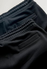 Pantaloni della tuta neri con vita elasticizzata e tasche posteriori. Tessuto morbido con una texture liscia, mostrato da una visuale dall'alto verso il basso.