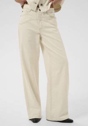 Personne portant un pantalon beige taille haute à jambes larges avec des coutures visibles et des chaussures noires pointues, mains ajustant l'ourlet d'une veste beige.