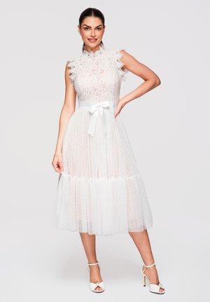 Femme souriante, portant une robe midi sans manches en dentelle blanche avec une ceinture en ruban de satin et des sandales blanches à talons hauts, posant la main sur la hanche.