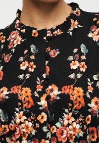 Μαύρη μπλούζα με πορτοκαλί και ροδακινί φλοράλ σχέδιο, γιακά με βολάν και κουμπιά καλυμμένα με ύφασμα κατά μήκος του μπροστινού μέρους.