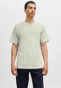 Tricou din bumbac gri deschis, cu guler rotund și mâneci scurte, având un logo discret pe piept și o croială lejeră. Purtați cu blugi închiși la culoare.
