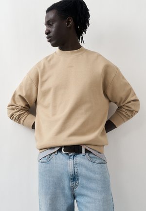 Νεαρό άτομο με σκούρο δέρμα και κάνναβους φοράει μπεζ φούτερ και ανοιχτό μπλε τζιν, στέκεται με τα χέρια πίσω από την πλάτη και κοιτάζει στο πλάι.