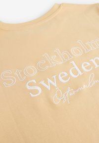 Vaalean keltainen puuvillapaita, jossa on valkoista brodeerattua tekstiä: "Tukholma, Ruotsi, Östermalm" vaihtelevilla fonttikokoilla ja -tyyleillä.