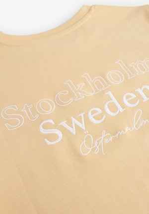 T-shirt in cotone giallo chiaro con testo ricamato bianco: "Stoccolma, Svezia, Östermalm" in diverse dimensioni e stili di carattere.