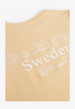 Camiseta de algodón amarillo claro con texto bordado en blanco: "Estocolmo, Suecia, Östermalm" en diferentes tamaños y estilos de fuente.