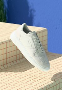 White sneaker featuring a mix of smooth leather and suede, textured perforations, flat laces, and a rubber sole, set against a geometric background.