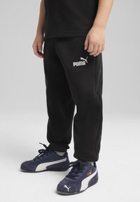 Juodos sportinės kelnės su laisvu kirpimu ir elastinga juosta, ant kairiojo kojos raudonai užrašytas baltoji "PUMA" logo. Suderintos su tamsiai mėlynomis sportinėmis bateliais.