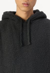 Sudadera con capucha de forro polar negro y superficie texturizada, que presenta un capuchón con cordones y costuras visibles para añadir un detalle adicional. Material suave y acogedor.