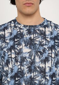 Mėlynos spalvos trumpomis rankovėmis T-shirt, puikuojantis delno medžio raštu šviesiame fone. Pagamintas iš medvilnės, su apvalia kaklo iškirpte ir laisvu kirpimu.