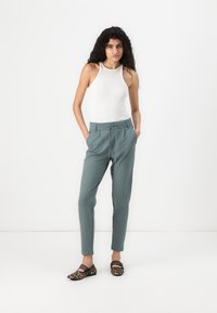 Lys grågrønne bukser med snor i livet, matchet med en tætsiddende hvid tanktop; modellen har mønstrede flade sko med sorte striber.
