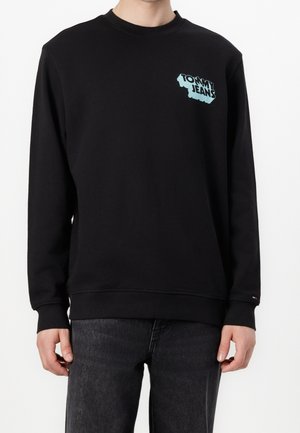 Czarna bluza z okrągłym dekoltem z jasnoniebieską grafiką "Tommy Jeans" na piersi, noszona przez osobę w ciemnych dżinsach.