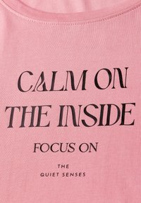 Růžové bavlněné tričko s krátkým rukávem, na kterém je výrazný černý text: "KLID UVNITŘ, SOUSTŘEĎTE SE NA TIŠE SMYSLY." Hladká textura.