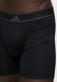 Gros plan d'une personne portant un short de sport noir ajusté Adidas avec un logo subtil à trois bandes sur la ceinture.