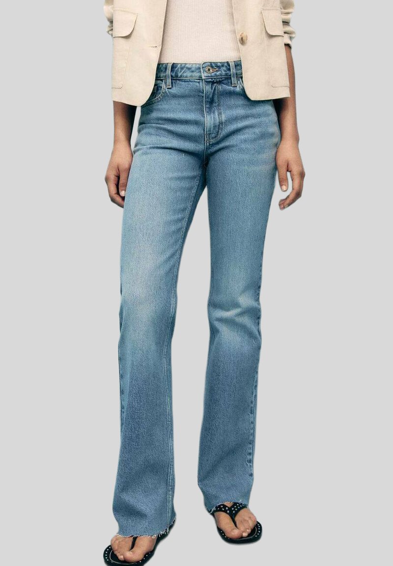 Jeans in denim azzurro chiaro a campana con un lavaggio sbiadito, vita a media altezza e design a cinque tasche. Indossati con sandali neri decorati con borchie.