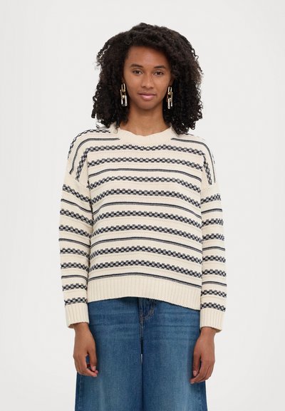 Maglione crema con righe navy, orlo e polsini a coste, con una vestibilità ampia e texture lavorata a maglia, abbinato a jeans in denim blu a gamba larga.