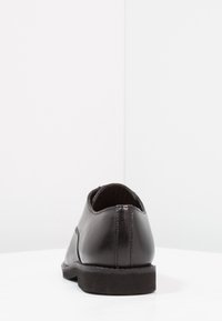 Black leather dress shoe viewed from the back, sitting on a white surface with a plain white background.
