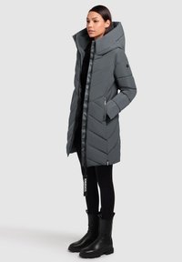 Grå dunjakke med chevron-syninger, høj krave, lynlås og to sidelommer, parret med sorte leggings og støvler.