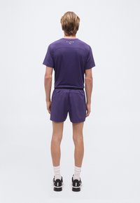 Άτομο στέκεται γυρισμένο προς τα πίσω φορώντας μια μοβ αθλητική στολή με σορτς, λευκές κάλτσες έως τον αστράγαλο και μαύρα παπούτσια Puma σε απλό φόντο.