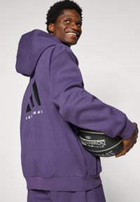 Paarse sweatvest met capuchon, met een groot zwart logo op de achterkant en geribbelde manchetten. De persoon houdt een basketbal vast.