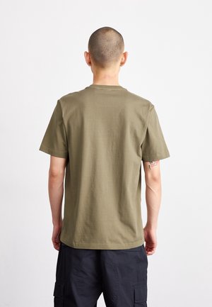Mand med barbert hoved, der vender væk, iført olivengrøn kortærmet t-shirt og sorte cargoshorts mod hvid baggrund.