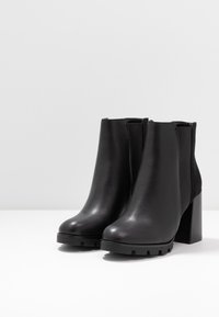 Musta nilkkurit, jotka on valmistettu sileästä nahasta ja joissa on sivulla joustavat paneelit, massiivinen korko ja kuvioitu pohja pitoon.