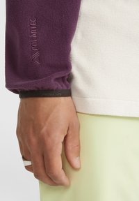 Giacca in fleece con manica viola e corpo bianco, caratterizzata da un tessuto testurizzato, polsini elasticizzati e un'etichetta di marca nera. Tessuto verde chiaro nella parte inferiore.