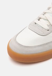 Bőr és nubuk sneaker fehér felsőrésszel, világosszürke részletekkel és gumis talppal. Jellemzői a lapos fűzők és a varrott részletek.