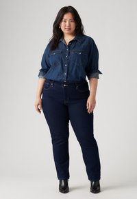 Mörkt tvättad denimskjorta med två bröstfickor, knäppning fram och uppvikta ärmar. Slim-fit mörka jeans och svarta ankelkängor med minimal detaljering.