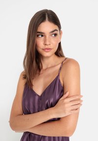 Fialová saténová košieľka s vertikálnymi pruhmi; má tenké ramienka a výstrih do V. Model má prirodzený vzhľad, rovné vlasy a minimálny make-up.