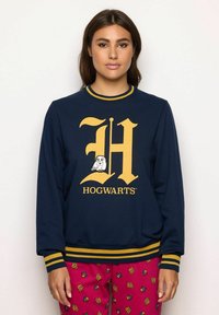 Donna che indossa una felpa blu navy con una grafica di un gufo e una "H" gialla, e pantaloni rossi con motivi ripetuti di "HP" e il cresti di una casa.