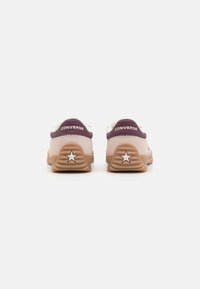 Converse-sko med en hvid ruskindsoverdel, mørk lilla hæl og beige gummisål med en stjernedetalje. Tekstureret med runde snørebånd.