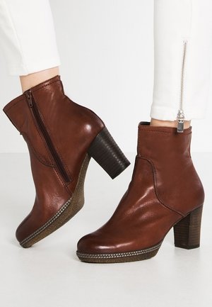 Bottines en cuir marron avec fermetures éclair latérales et talons blocs empilés, portées avec un pantalon blanc avec fermetures éclair aux chevilles.