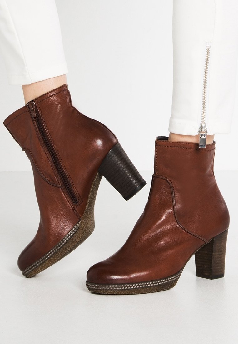 Bottines en cuir marron avec fermetures éclair latérales et talons blocs empilés, portées avec un pantalon blanc avec fermetures éclair aux chevilles.