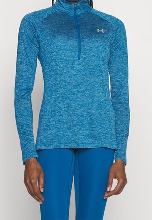 Person trägt ein blaues, langärmeliges Sportoberteil mit einem Viertelreißverschluss und passenden blauen Leggings, steht vor einem einfarbigen Hintergrund.