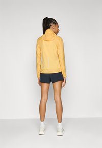 Veste légère jaune avec capuche, dotée d'une fermeture éclair à l'avant et d'un détail de logo réfléchissant. Associée à un short noir et des baskets de sport.