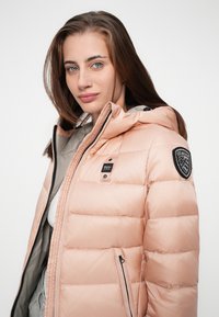Perzikkleurige pufferjack met een capuchon, voorzien van een ritssluiting aan de voorkant, zijzakken en een logopatch op de linkerschouder. Gladde, gewatteerde textuur.