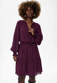 Paarse jurk met lange mouwen en een V-hals, elastische tailleband en gerimpelde manchetten. Heeft een gelaagde rok en een subtiel stippenpatroon.