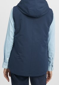 Mörkblå vadderad väst med huva, med en slät textur och skräddarsydd passform, bärs över en ljusblå långärmad skjorta.