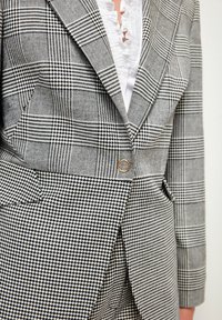 Mustavalkoinen ruudullinen bleiseri, jossa on yhdistelmä houndstooth- ja ruutukuosisia, jossa on yksittäinen nappi ja terävät läpät. Rakenteellinen kangas.