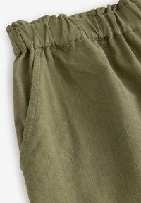 Pantalon en lin vert olive avec taille élastique et coutures visibles sur la poche latérale, montrant la texture du tissu et un détail froncé à la taille.