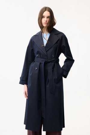 Ung kvinde med langt brunt hår iført en navyblå trenchcoat med bælte over en lyseblå top, stående mod en ensfarvet hvid baggrund.