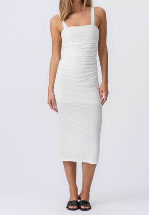 Kobieta ubrana w białą, marszczoną, bezrękawną sukienkę midi z kwadratowym dekoltem oraz czarne, odkryte sandały na płaskiej podeszwie, stojąca na tle jednolitego tła.