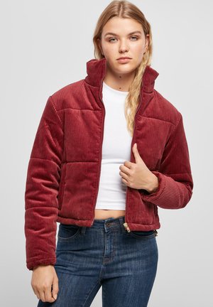 Rode corduroy jas met een hoge kraag, ritssluiting aan de voorkant en een patroon van gestikte delen. Gepaard met een witte top en blauwe spijkerbroek.