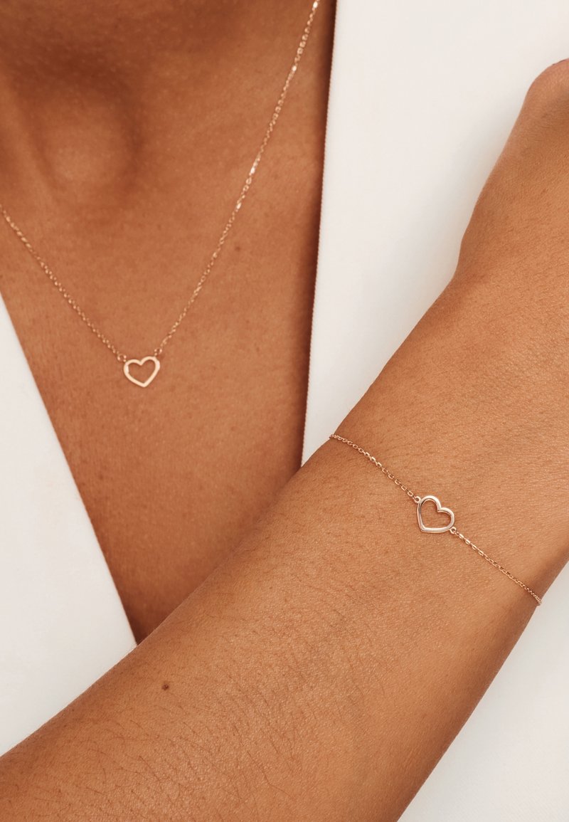 Roze gouden hart hanger ketting en armband, beide met delicate kettingen en open hart vormen, tentoongesteld op een huidkleurige arm.